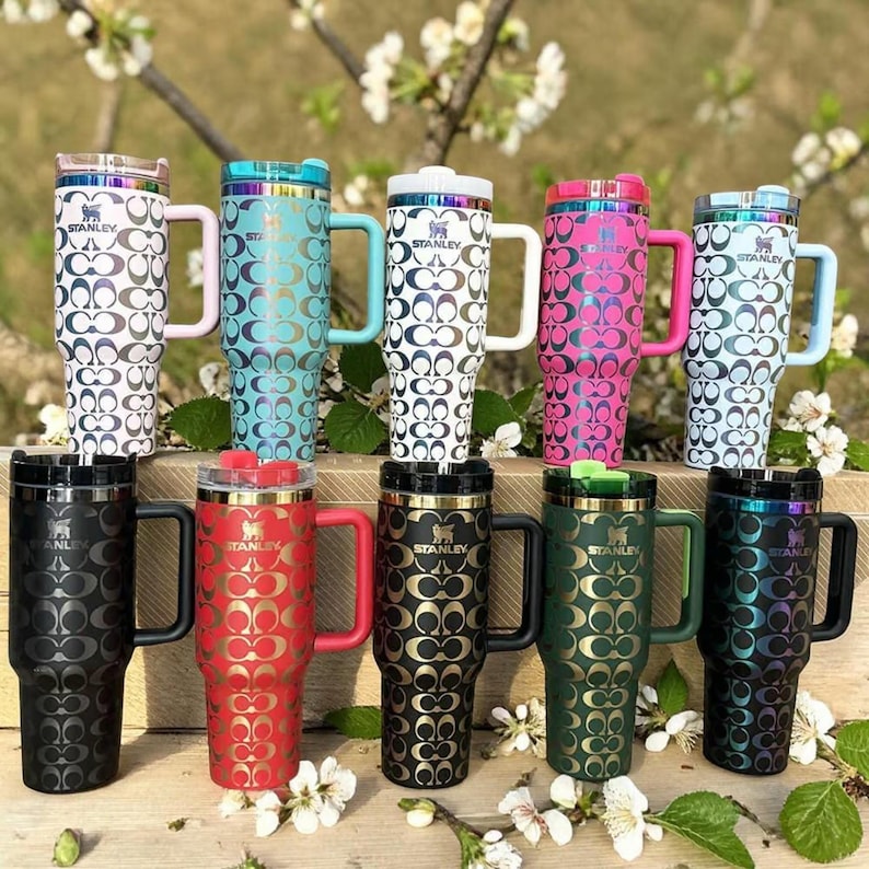 May include: Ten Stanley tumblers with handles and lids, arranged in two rows. Each tumbler features a repeating interlocking C logo pattern. The tumblers come in various colors including white, teal, pink, black, red, green, and iridescent finishes.
