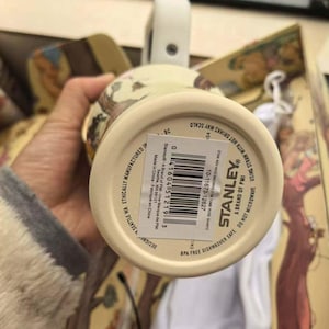 May include: A cream-colored Stanley mug with a white handle and a floral design. The bottom of the mug has a label with the Stanley logo and product information. The mug is BPA-free and dishwasher safe.