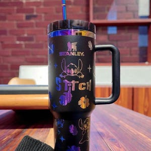 K&ouml;nnte beinhalten: Schwarzer Stanley-Becher mit Griff und Stitch-Design. Der Becher hat einen schwarzen Deckel, einen Strohhalm und schillernde Akzente. Die Texte "STANLEY" und "Stitch" sind auf dem Becher sichtbar.