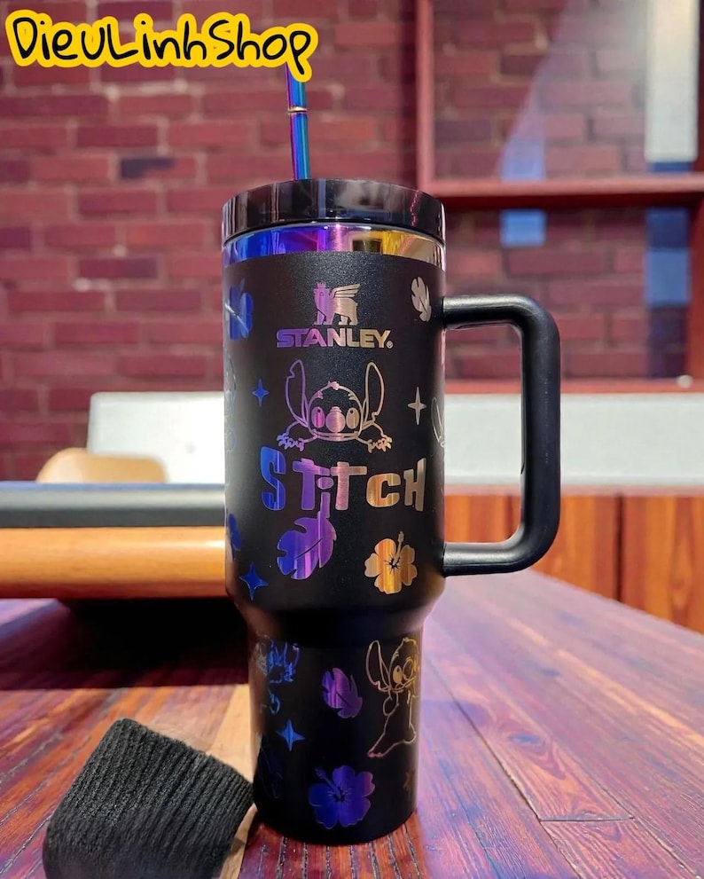 K&ouml;nnte beinhalten: Schwarzer Stanley-Becher mit Griff und Stitch-Design. Der Becher hat einen schwarzen Deckel, einen Strohhalm und schillernde Akzente. Die Texte "STANLEY" und "Stitch" sind auf dem Becher sichtbar.