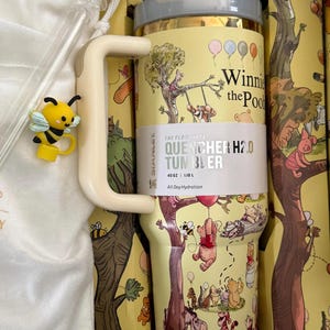 May include: A yellow Stanley Quencher H2.0 tumbler featuring Winnie the Pooh characters and illustrations. The tumbler has a handle, a straw, and a lid. A yellow bee charm is attached to the handle. The tumbler holds 40 oz (1.18 L) and is designed for all-day hydration.