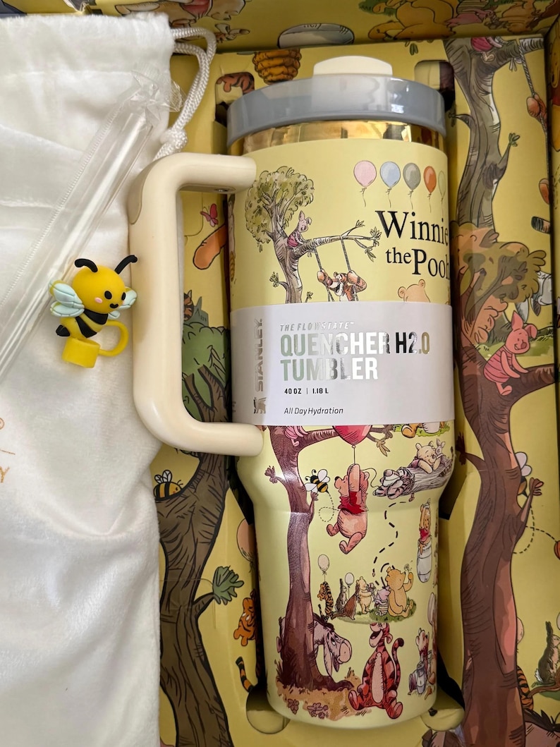 May include: A yellow Stanley Quencher H2.0 tumbler featuring Winnie the Pooh characters and illustrations. The tumbler has a handle, a straw, and a lid. A yellow bee charm is attached to the handle. The tumbler holds 40 oz (1.18 L) and is designed for all-day hydration.