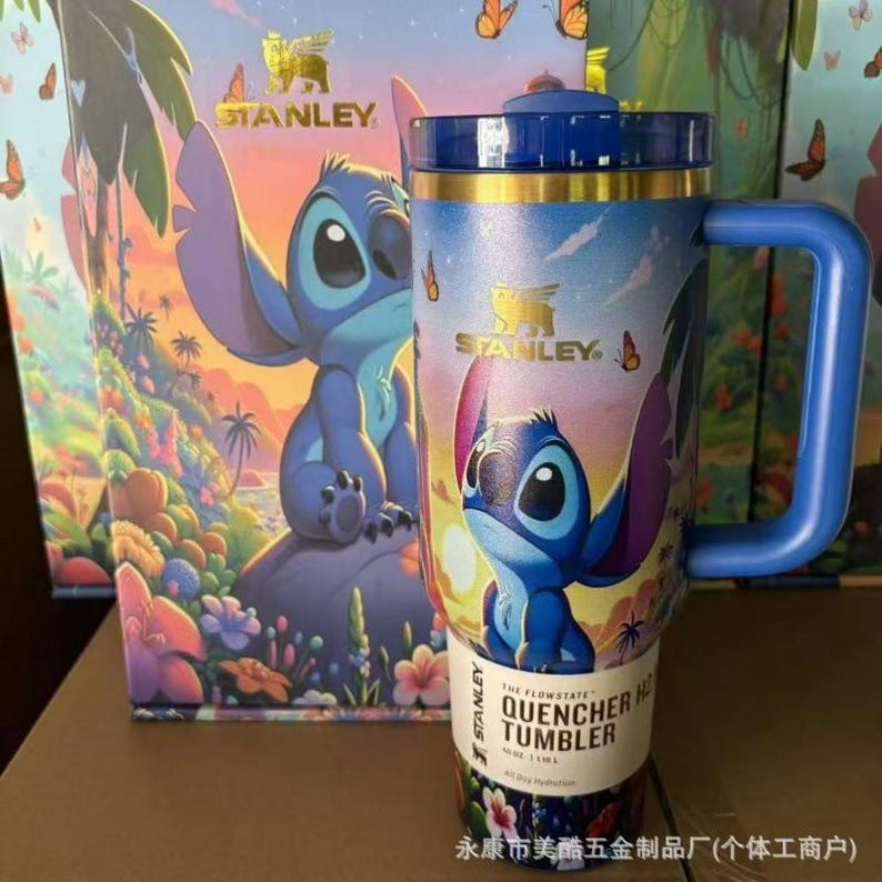 May include: A blue Stanley Flowstate Quencher H2.0 tumbler featuring a graphic of the character Stitch from Lilo & Stitch, with a tropical floral background, a blue handle, a blue lid, and a gold-coloured rim, standing in front of its matching box.