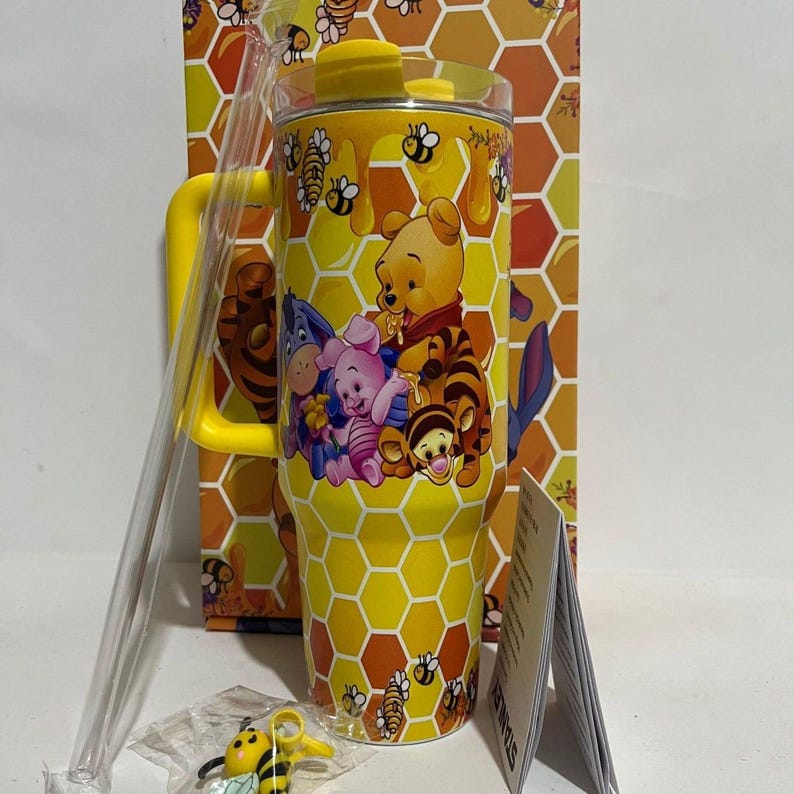 May include: Yellow tumbler featuring Winnie the Pooh and friends, with a honeycomb pattern and bee accents. Includes a yellow handle, lid, and straw. A small bee charm and packaging are also visible.