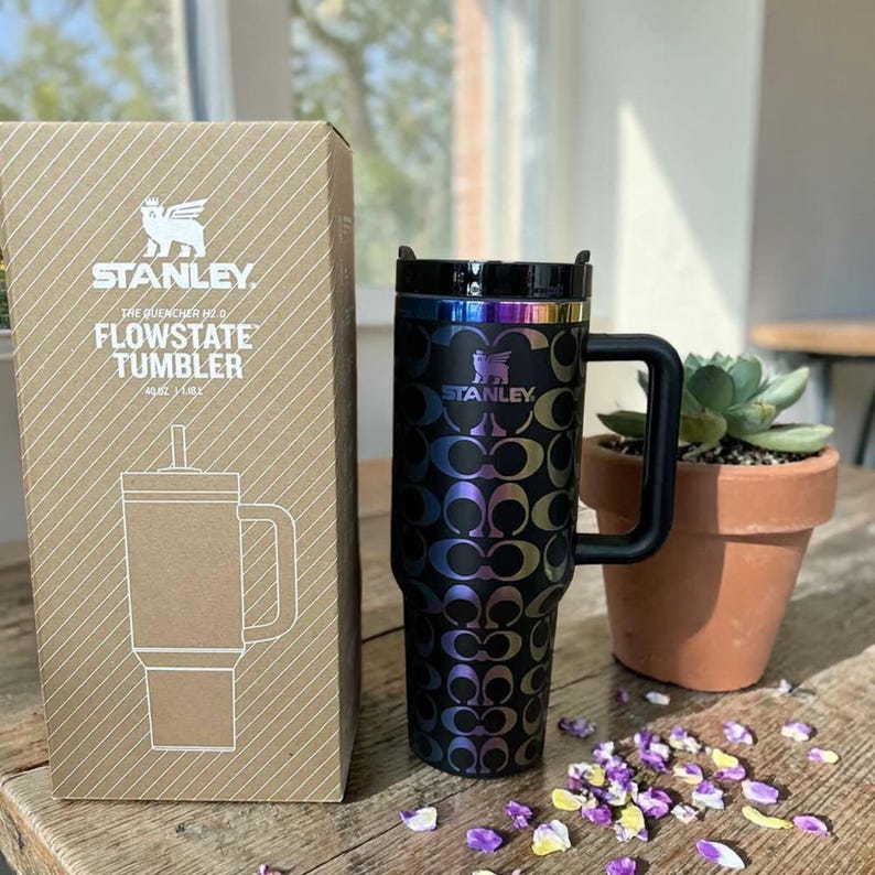 May include: A black Stanley Flowstate tumbler with a repeating iridescent C-pattern, a black handle, and a black lid. The tumbler is next to its cardboard packaging, which features the Stanley logo and the text The Quencher H2.0 Flowstate Tumbler.