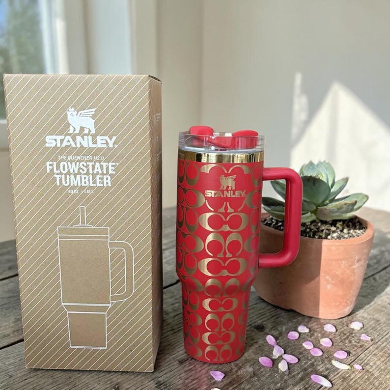 May include: A red Stanley Flowstate tumbler with a gold repeating logo pattern, a red handle, and a clear lid with a red straw opening, standing next to its cardboard box labeled Stanley The Quencher H2.0 Flowstate Tumbler 40 oz 1.18 L.