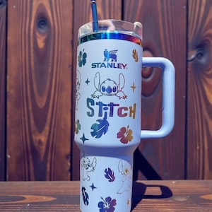 K&ouml;nnte beinhalten: Ein hellblauer Stanley-Becher mit Griff und Stitch-Design. Der Becher hat einen Strohhalm und einen transparenten Deckel mit einem regenbogenfarbenen, schillernden Rand. Das Design umfasst das Wort "Stitch" und verschiedene Blumen- und Sterngrafiken.