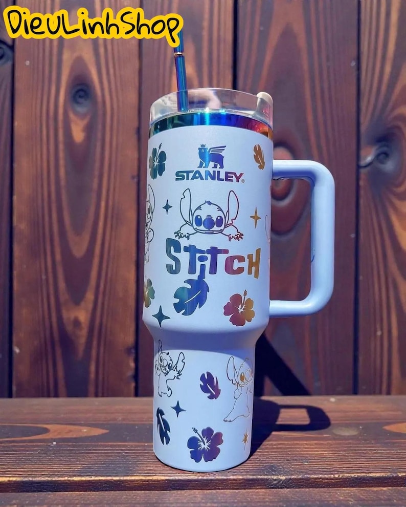 K&ouml;nnte beinhalten: Ein hellblauer Stanley-Becher mit Griff und Stitch-Design. Der Becher hat einen Strohhalm und einen transparenten Deckel mit einem regenbogenfarbenen, schillernden Rand. Das Design umfasst das Wort "Stitch" und verschiedene Blumen- und Sterngrafiken.
