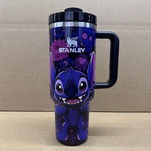 May include: A dark blue Stanley tumbler featuring a graphic of the character Stitch with large ears and a wide smile. The design includes purple and pink floral and skull patterns, a black handle, and a black lid with the Stanley logo printed in white.