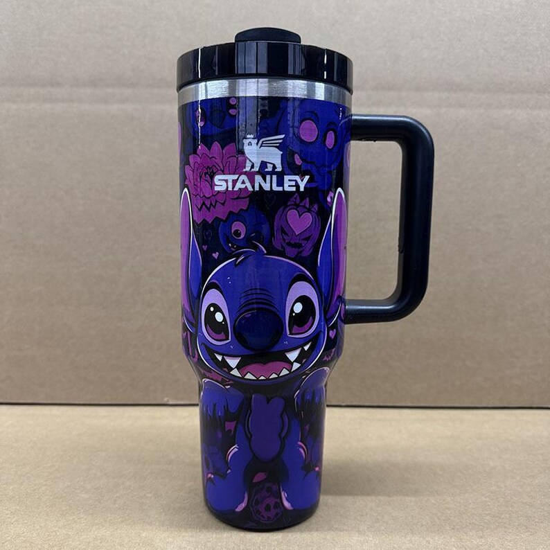 May include: A dark blue Stanley tumbler featuring a graphic of the character Stitch with large ears and a wide smile. The design includes purple and pink floral and skull patterns, a black handle, and a black lid with the Stanley logo printed in white.