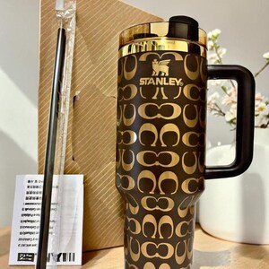 May include: A dark brown Stanley tumbler featuring a repeating gold-toned logo pattern, a black handle, and a gold-rimmed lid. A black straw in a clear plastic sleeve and a white instruction manual are positioned next to the tumbler.