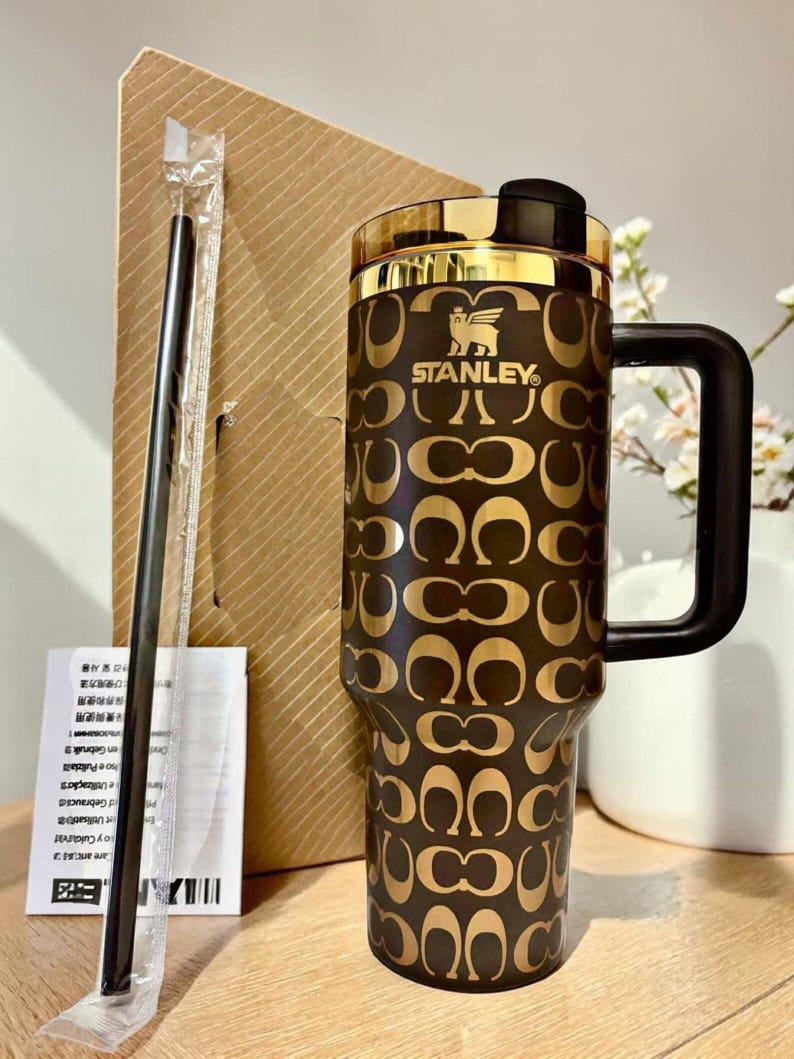May include: A dark brown Stanley tumbler featuring a repeating gold-toned logo pattern, a black handle, and a gold-rimmed lid. A black straw in a clear plastic sleeve and a white instruction manual are positioned next to the tumbler.