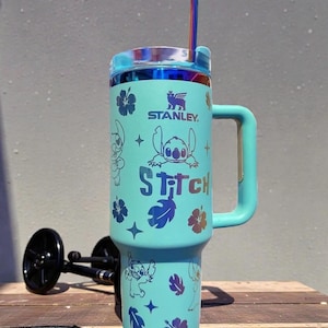 K&ouml;nnte beinhalten: Ein t&uuml;rkisfarbener Stanley-Becher mit Stitch-Grafiken, Blumen und Sternen. Der Becher hat einen Griff, einen Deckel und einen Strohhalm. Der Text "Stitch" ist auf der Vorderseite aufgedruckt. Der Text "DieuLinhShop" befindet sich oben links.