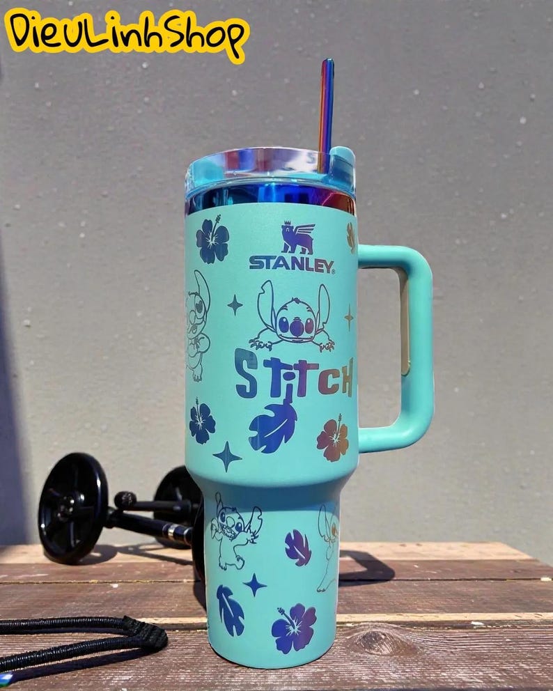 K&ouml;nnte beinhalten: Ein t&uuml;rkisfarbener Stanley-Becher mit Stitch-Grafiken, Blumen und Sternen. Der Becher hat einen Griff, einen Deckel und einen Strohhalm. Der Text "Stitch" ist auf der Vorderseite aufgedruckt. Der Text "DieuLinhShop" befindet sich oben links.