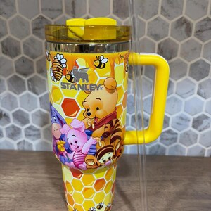 May include: A yellow and stainless steel Stanley tumbler featuring a Winnie the Pooh design. The tumbler has a yellow handle, lid, and a clear straw. The design includes Winnie the Pooh, Piglet, Eeyore, and Tigger characters, with a honeycomb and bee pattern.