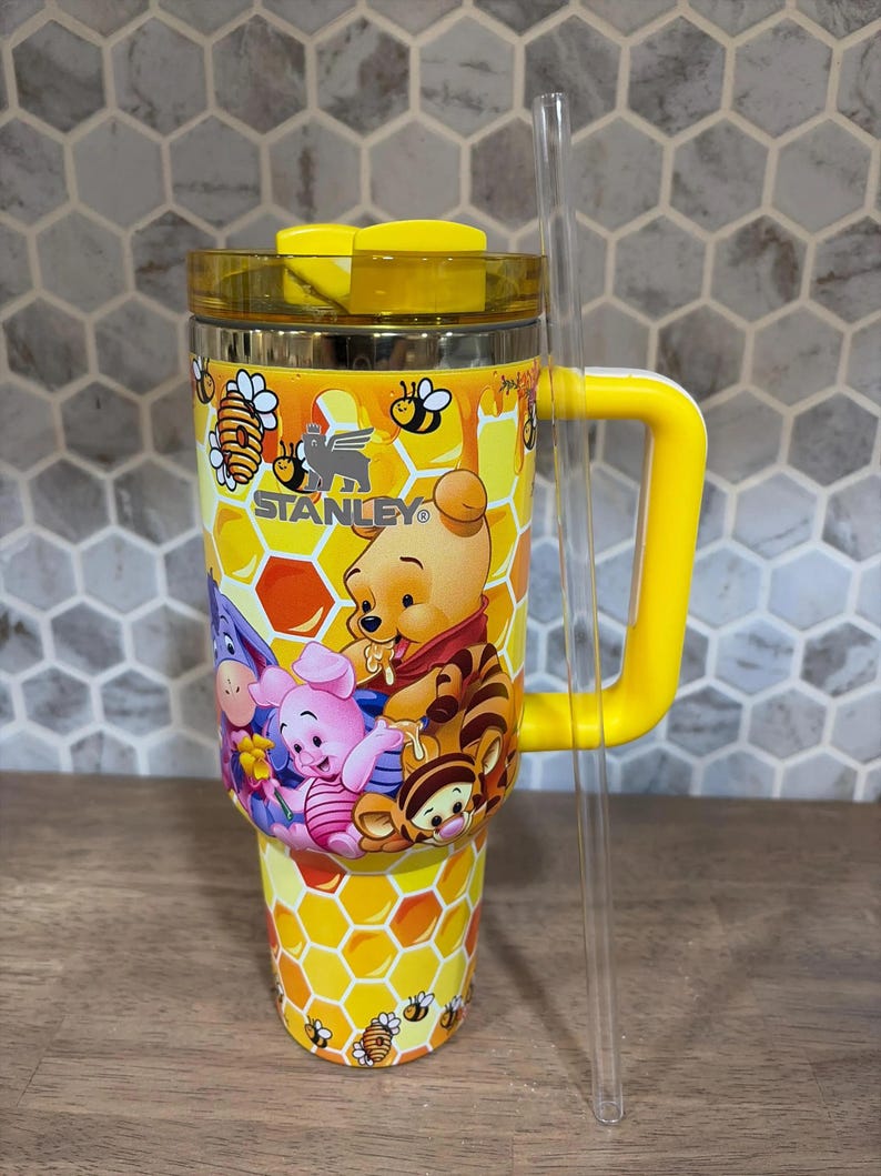 May include: A yellow and stainless steel Stanley tumbler featuring a Winnie the Pooh design. The tumbler has a yellow handle, lid, and a clear straw. The design includes Winnie the Pooh, Piglet, Eeyore, and Tigger characters, with a honeycomb and bee pattern.