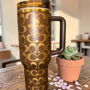 May include: A brown Stanley tumbler with a gold repeating C-logo pattern, a dark brown handle, and a translucent brown lid. The front features the gold Stanley logo with the text "STANLEY SINCE 1913" and a winged bear icon centered on the upper body.