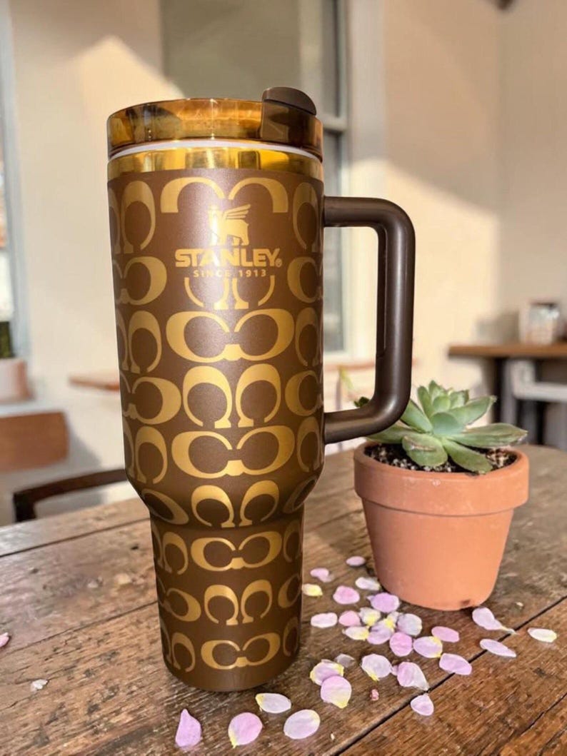 May include: A brown Stanley tumbler with a gold repeating C-logo pattern, a dark brown handle, and a translucent brown lid. The front features the gold Stanley logo with the text "STANLEY SINCE 1913" and a winged bear icon centered on the upper body.