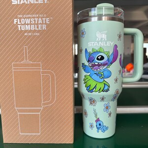 May include: A light green Stanley Quencher H2.0 FlowState tumbler with a 40 oz capacity, featuring a graphic of the character Stitch in a hula skirt surrounded by small blue and white flowers, next to its cardboard packaging box with white line art.