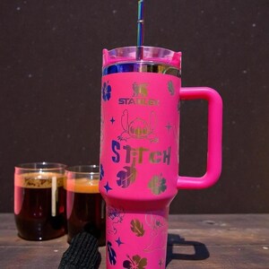 K&ouml;nnte beinhalten: Ein pinkfarbener Stanley-Becher mit Griff, mit einem Design der Figur Stitch und dem Wort "Stitch". Der Becher hat einen Deckel und einen regenbogenfarbenen Strohhalm. Im Hintergrund sind zwei Gl&auml;ser mit einem dunklen Getr&auml;nk.