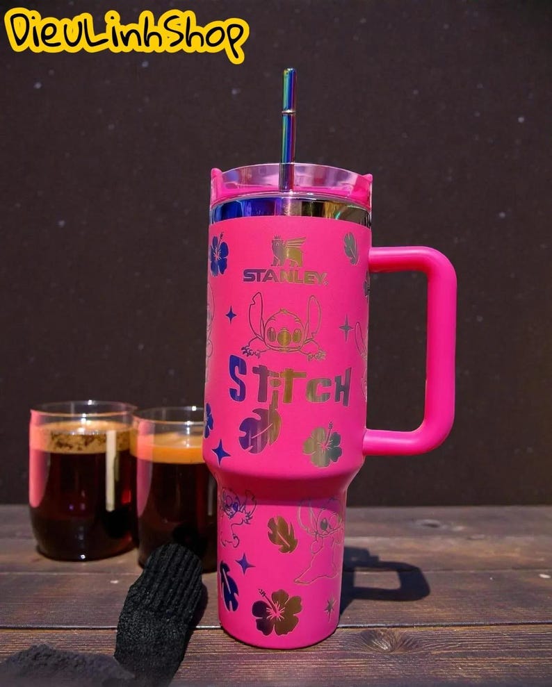 K&ouml;nnte beinhalten: Ein pinkfarbener Stanley-Becher mit Griff, mit einem Design der Figur Stitch und dem Wort "Stitch". Der Becher hat einen Deckel und einen regenbogenfarbenen Strohhalm. Im Hintergrund sind zwei Gl&auml;ser mit einem dunklen Getr&auml;nk.