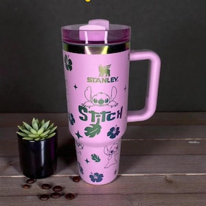 K&ouml;nnte beinhalten: Ein pinkfarbener Stanley-Reisebecher mit Griff und transparentem Deckel, der die Figur Stitch aus Lilo & Stitch zeigt. Das Design umfasst Stitch, Blumen und das Wort "Stitch". Ein kleiner Topf mit Pflanze und Kaffeebohnen befinden sich im Hintergrund.