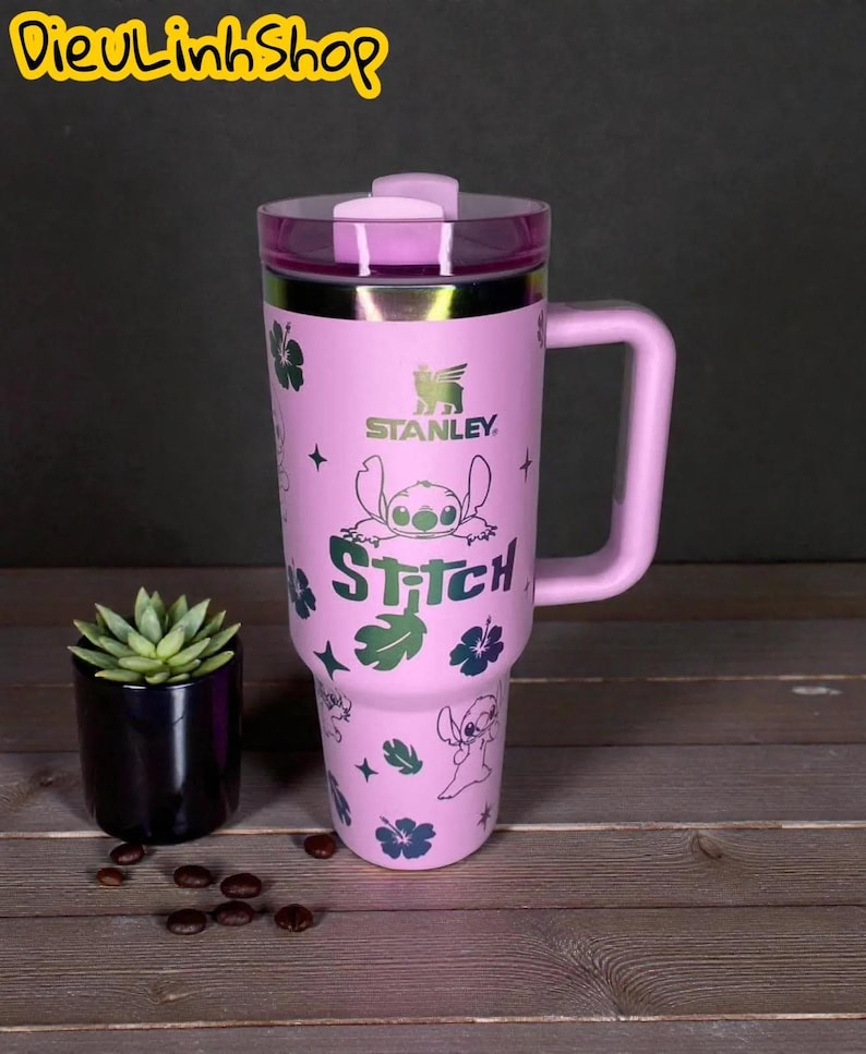 K&ouml;nnte beinhalten: Ein pinkfarbener Stanley-Reisebecher mit Griff und transparentem Deckel, der die Figur Stitch aus Lilo & Stitch zeigt. Das Design umfasst Stitch, Blumen und das Wort "Stitch". Ein kleiner Topf mit Pflanze und Kaffeebohnen befinden sich im Hintergrund.