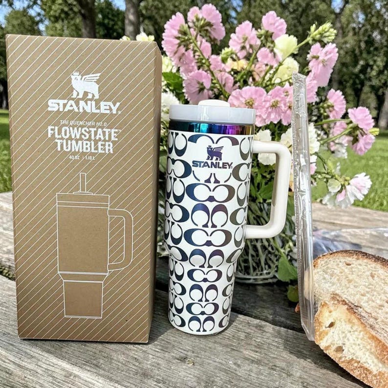 May include: A Stanley Flowstate 40 oz tumbler with a white and dark blue repeating logo pattern, a white handle, and a metallic rim, standing next to its brown cardboard packaging box which displays the Stanley logo and product name text.