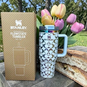 May include: A white Stanley Flowstate tumbler with a repeating metallic C-logo pattern, a handle, and a clear lid, standing next to its brown cardboard box labeled with the product name and 40oz capacity, set against a background of tulips.
