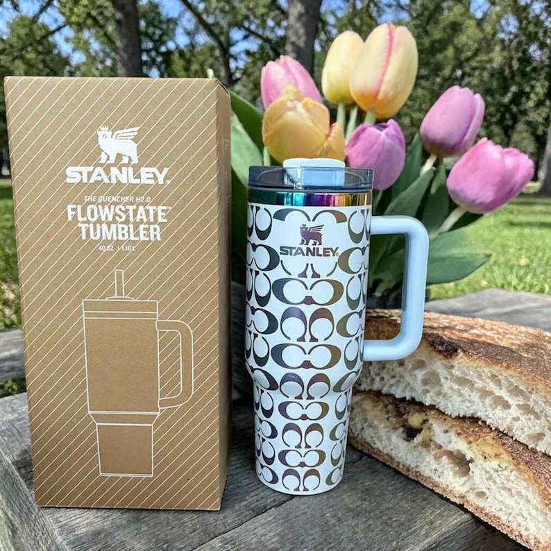 May include: A white Stanley Flowstate tumbler with a repeating metallic C-logo pattern, a handle, and a clear lid, standing next to its brown cardboard box labeled with the product name and 40oz capacity, set against a background of tulips.