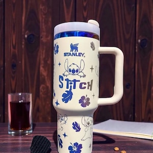 K&ouml;nnte beinhalten: Wei&szlig;er Stanley-Reisebecher mit Stitch aus Lilo & Stitch. Der Becher hat einen wei&szlig;en Griff und Deckel mit blau-violettem Rand. Der Becher ist mit Stitch-Illustrationen, blauen Blumen und dem Wort "Stitch" verziert.