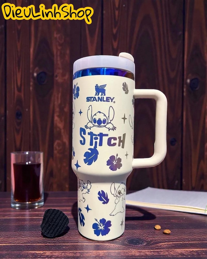 K&ouml;nnte beinhalten: Wei&szlig;er Stanley-Reisebecher mit Stitch aus Lilo & Stitch. Der Becher hat einen wei&szlig;en Griff und Deckel mit blau-violettem Rand. Der Becher ist mit Stitch-Illustrationen, blauen Blumen und dem Wort "Stitch" verziert.