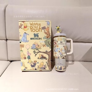 May include: A Winnie the Pooh themed Stanley travel mug and matching gift box. The mug features a white handle, a yellow lid with a bee, and illustrations of Pooh and friends. The box has similar illustrations.