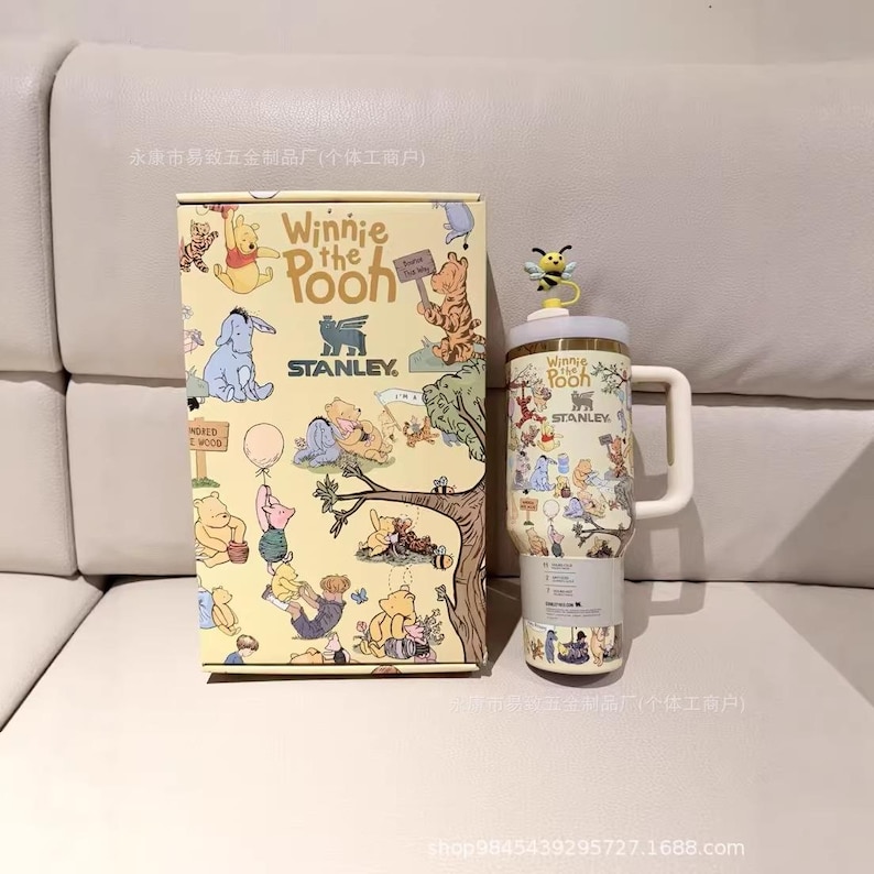 May include: A Winnie the Pooh themed Stanley travel mug and matching gift box. The mug features a white handle, a yellow lid with a bee, and illustrations of Pooh and friends. The box has similar illustrations.