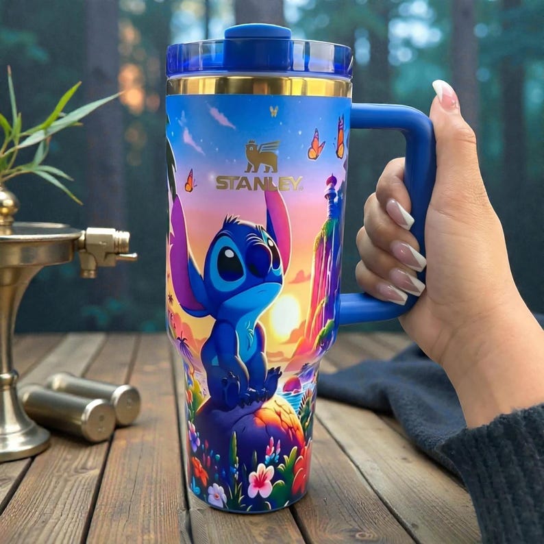 May include: A blue Stanley tumbler featuring a colourful graphic of the character Stitch sitting on a rock, surrounded by tropical flowers, butterflies, and a sunset landscape. The cup has a blue handle, a gold-tone rim, and a clear blue lid.
