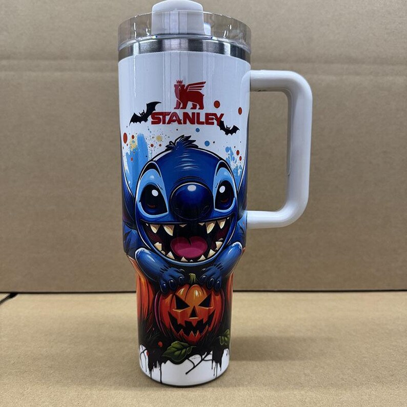 May include: A white stainless steel tumbler with a handle and a clear lid, featuring a graphic of a blue alien character above a carved orange jack-o'-lantern pumpkin. The Stanley logo is printed in red near the top with black bat silhouettes.