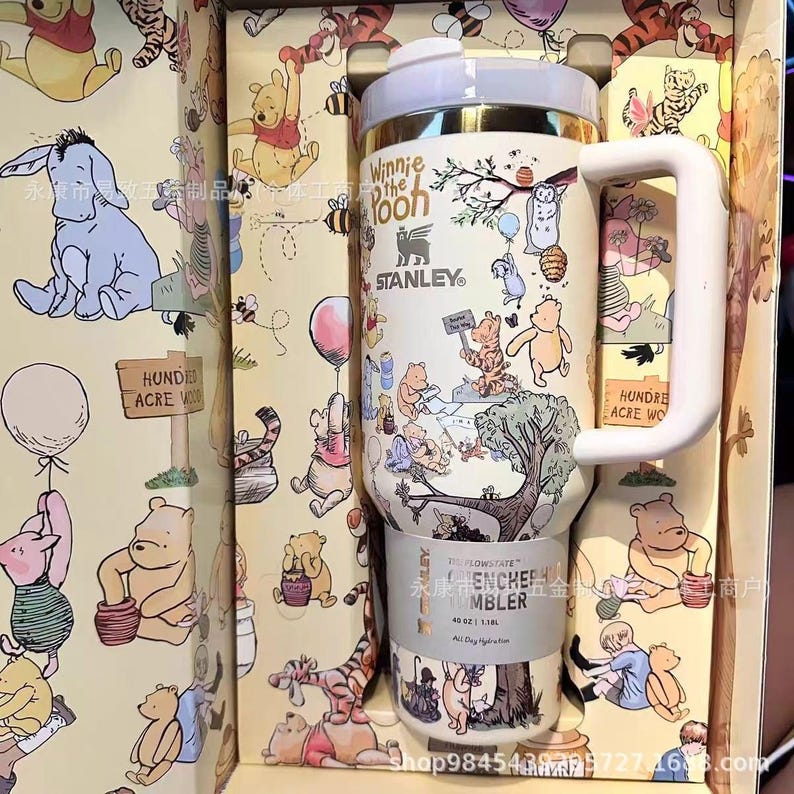 May include: A Winnie the Pooh themed Stanley tumbler with a white handle and lid. The tumbler features illustrations of Winnie the Pooh and friends, with the words "Winnie the Pooh" and "Stanley" visible. The tumbler is in a decorative box.