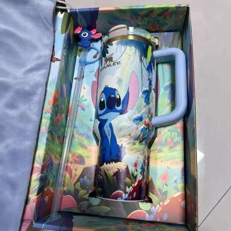 May include: A stainless steel tumbler with a handle and a matching straw, featuring a blue character with large ears and a tropical forest print. The Stanley brand name is printed on the upper portion of the tumbler, which is housed in a decorative box.