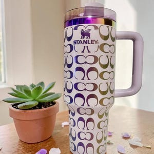 May include: A white Stanley tumbler with a handle and a purple iridescent lid. The body features a repeating pattern of metallic, color-shifting C-shaped logos and the Stanley brand name with a winged bear logo printed in dark purple on the upper front.