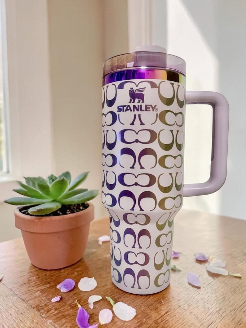 May include: A white Stanley tumbler with a handle and a purple iridescent lid. The body features a repeating pattern of metallic, color-shifting C-shaped logos and the Stanley brand name with a winged bear logo printed in dark purple on the upper front.