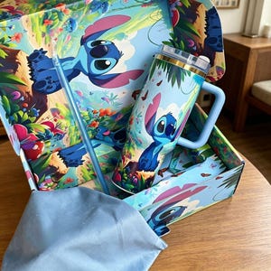 May include: A light blue tumbler with a handle, a clear lid, and a matching straw, featuring a graphic of the blue alien character Stitch in a tropical setting. The tumbler is presented inside a matching decorative box with a blue cloth.