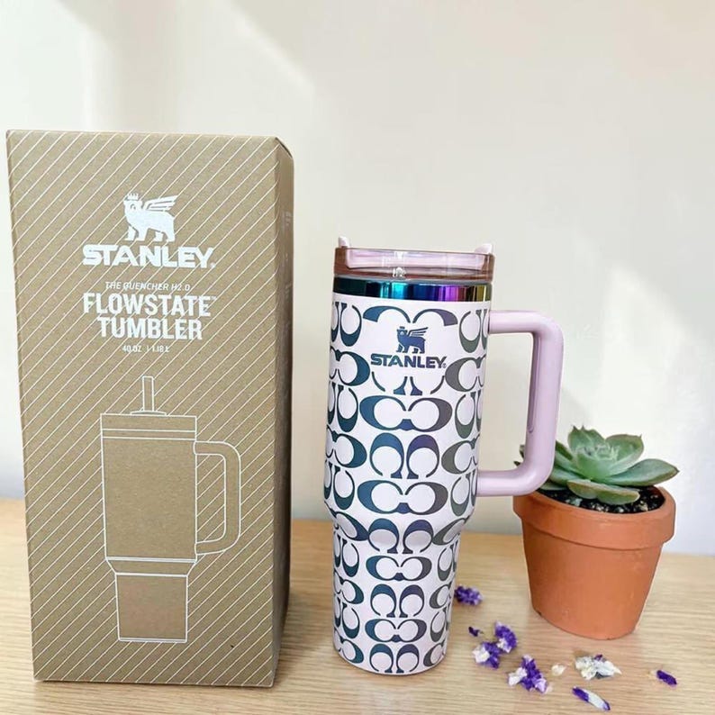 May include: A 40 oz Stanley Flowstate tumbler with a repeating dark logo pattern on a light background, featuring a light pink handle, a metallic rainbow rim, and a clear lid. The tumbler is displayed next to its brown cardboard packaging box.
