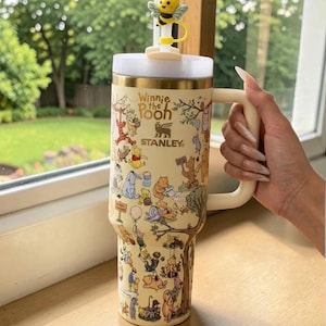 May include: A beige Stanley tumbler featuring Winnie the Pooh characters and the text "Winnie the Pooh". The tumbler has a white lid, a gold rim, and a cream-colored handle. A yellow bee-shaped topper sits on the lid.
