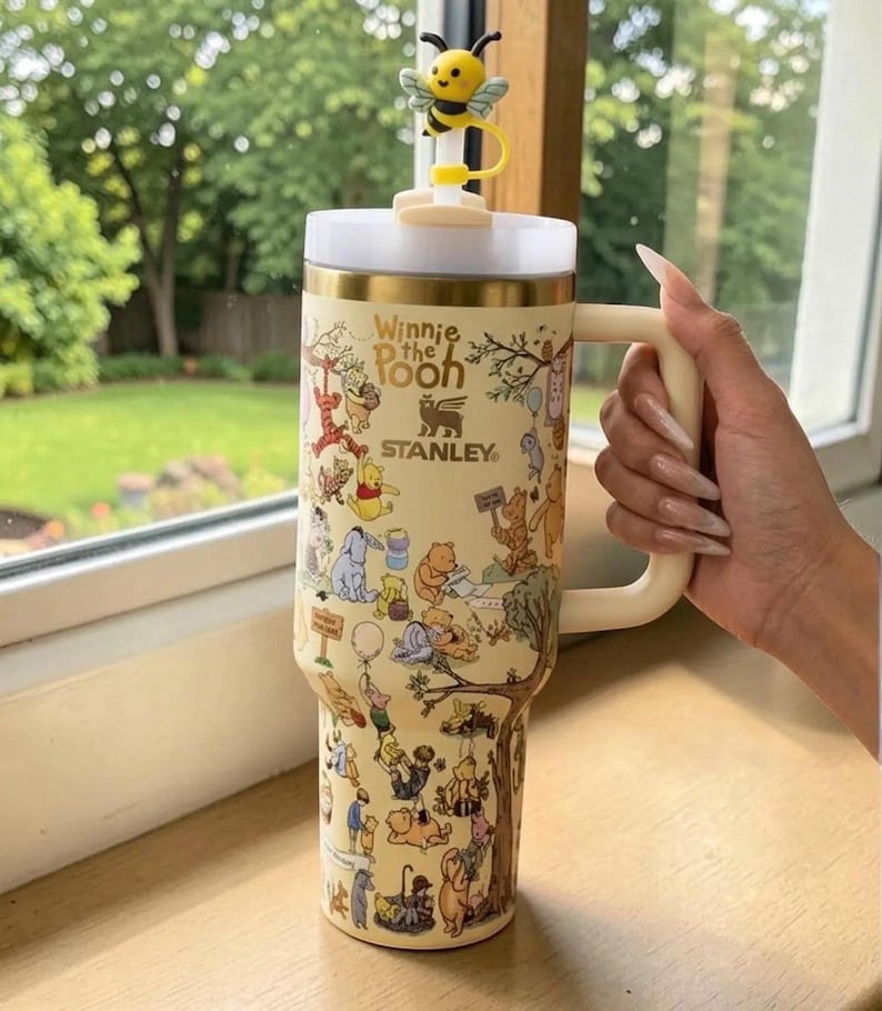 May include: A beige Stanley tumbler featuring Winnie the Pooh characters and the text "Winnie the Pooh". The tumbler has a white lid, a gold rim, and a cream-colored handle. A yellow bee-shaped topper sits on the lid.