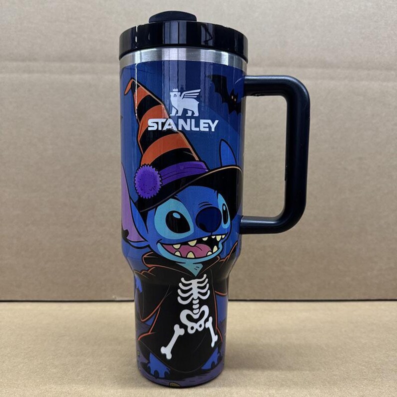 May include: A blue Stanley tumbler with a black lid and handle featuring a graphic of the character Stitch wearing a black and orange striped witch hat and a black skeleton costume. The design includes a small bat silhouette and the Stanley logo.
