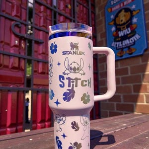 K&ouml;nnte beinhalten: Ein wei&szlig;er Stanley-Becher mit Griff und transparentem Deckel mit blauem Rand. Der Becher ist mit Illustrationen von Stitch, Hibiskusbl&uuml;ten und dem Wort "Stitch" verziert. Ein praktischer Begleiter f&uuml;r unterwegs.