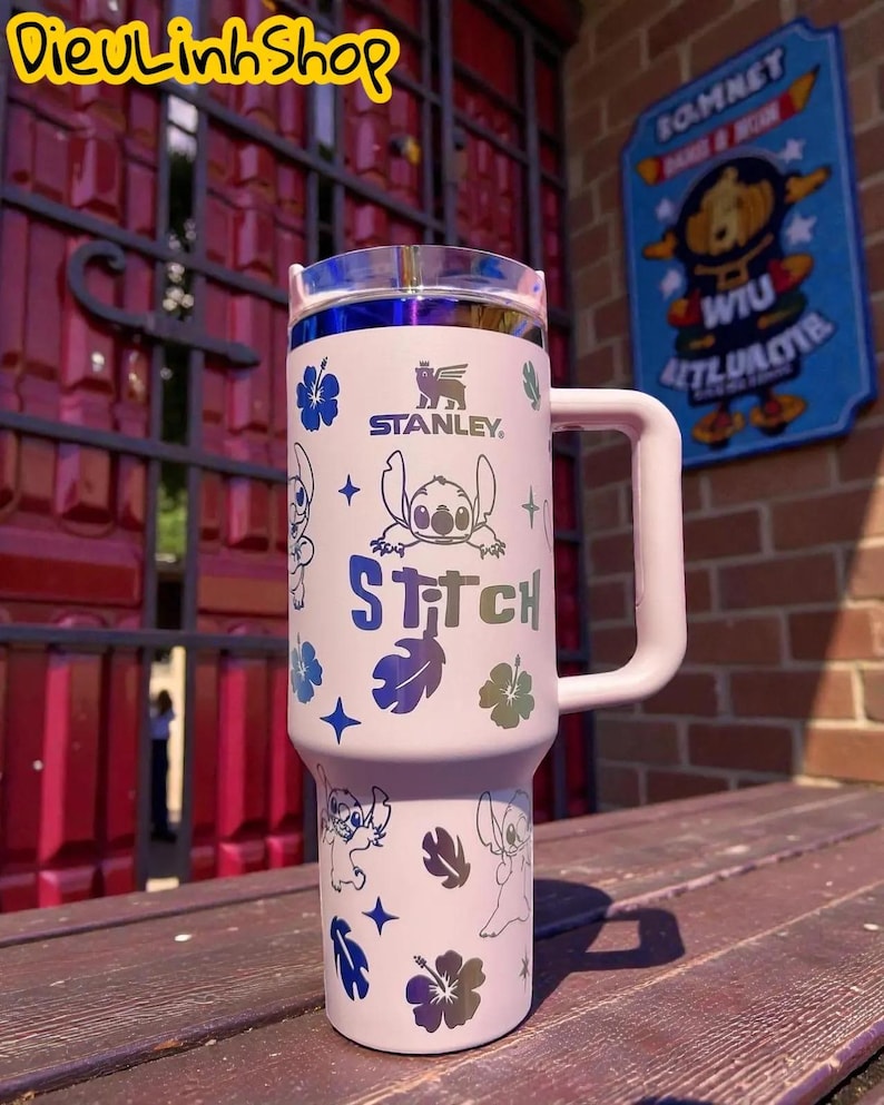 K&ouml;nnte beinhalten: Ein wei&szlig;er Stanley-Becher mit Griff und transparentem Deckel mit blauem Rand. Der Becher ist mit Illustrationen von Stitch, Hibiskusbl&uuml;ten und dem Wort "Stitch" verziert. Ein praktischer Begleiter f&uuml;r unterwegs.