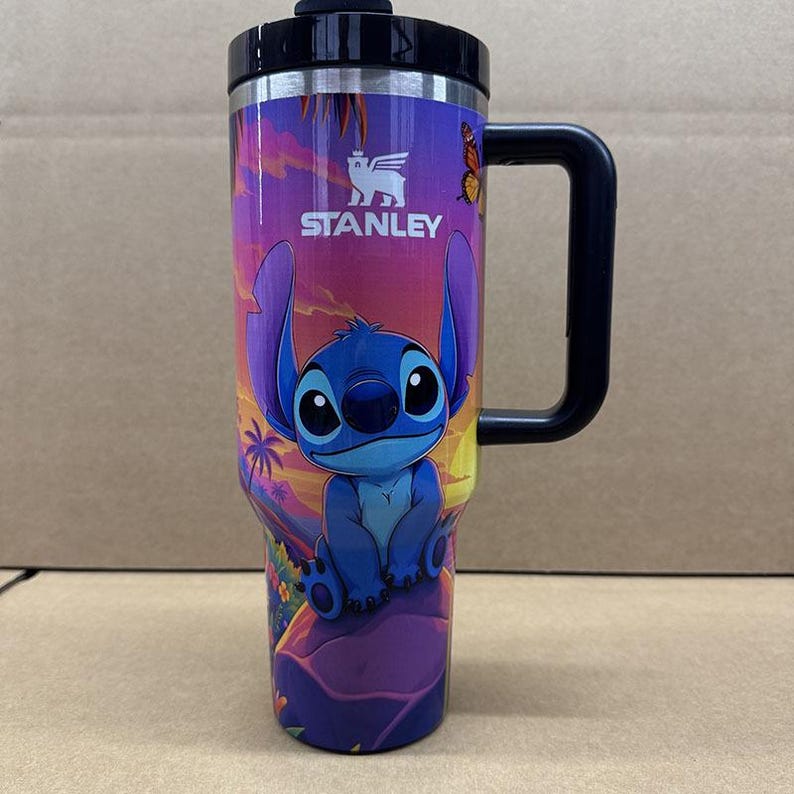 May include: A Stanley brand tumbler with a black handle and lid, featuring a colourful graphic of the character Stitch sitting on a rock. The background displays a sunset scene with palm trees, flowers, and a butterfly, all printed on the exterior.