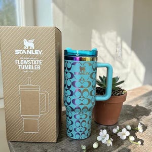 May include: A light blue Stanley Flowstate tumbler with a 40 oz capacity, featuring a repeating dark metallic logo pattern, a matching handle, and a translucent lid. The tumbler is displayed next to its original cardboard packaging box.