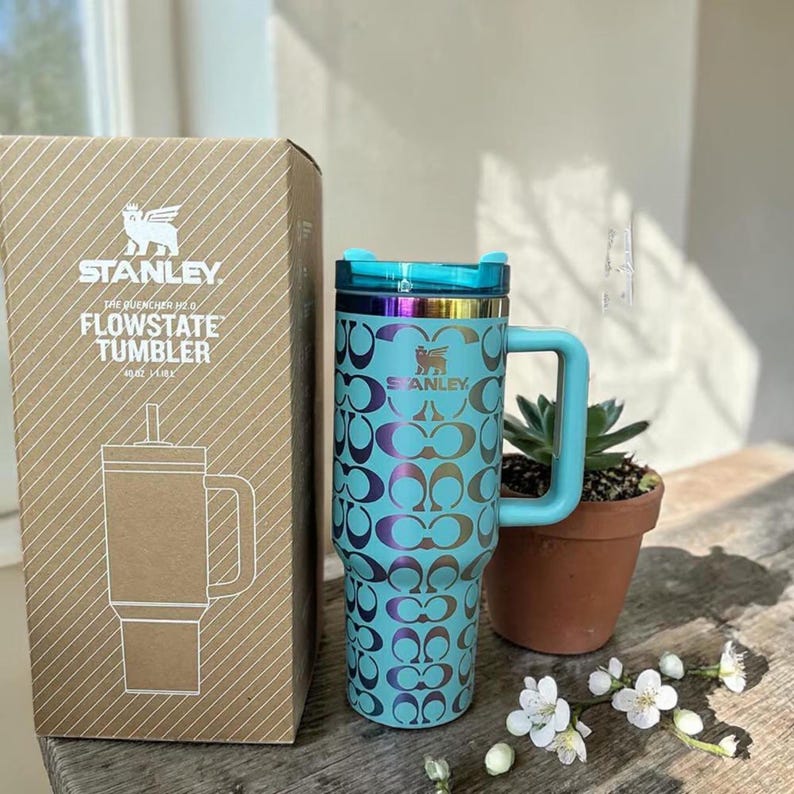 May include: A light blue Stanley Flowstate tumbler with a 40 oz capacity, featuring a repeating dark metallic logo pattern, a matching handle, and a translucent lid. The tumbler is displayed next to its original cardboard packaging box.